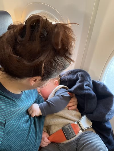 An adult and a baby are seated inside an aircraft cabin, presumably in or near Lautzenhausen, Germany, given the presence of an aircraft window. The adult, with brown hair styled in a messy bun secured by a dark claw clip, is visible from the back of the head and left shoulder, wearing a teal and dark blue striped long-sleeve shirt. A small silver stud earring is present in the adult's left ear. The baby, dressed in a dark long-sleeve top under a light brown sleeveless vest and dark grey corduroy pants, is resting against the adult's chest, with a flushed cheek visible. The baby's left hand is curled into a fist against the adult's shirt. An orange aircraft seatbelt with a silver buckle is secured around the baby. A dark blue blanket or garment is draped over the baby's right side. Two circular aircraft windows are visible in the background, showing diffused outdoor light.