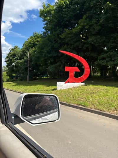 A red hammer and sickle sculpture is visible on a grassy mound next to a road. The sculpture, a symbol of communism, is large and metallic, with the hammer and sickle intertwined. The base of the sculpture is white and appears to be concrete. The road is asphalt and runs alongside the grassy area. In the foreground, a car's side mirror is visible, reflecting a person's hand holding a phone. The mirror has text that reads "OBJECTS IN MIRROR ARE CLOSER THAN THEY APPEAR". The background consists of lush green trees and a blue sky with white clouds. The scene appears to be in Pereslavl-Zalessky, Russia, a city known for its historical significance and Soviet-era monuments.