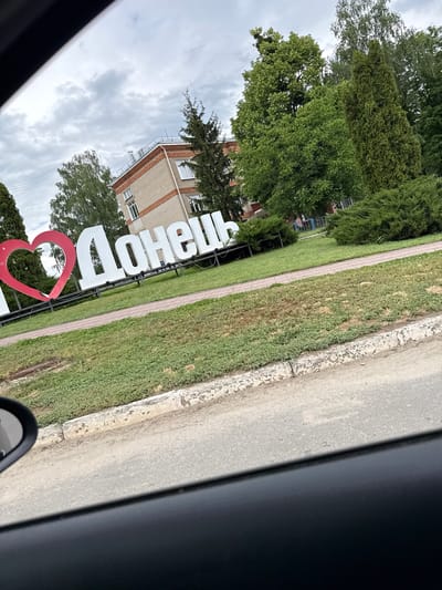 The image, captured from the perspective of a vehicle, displays an outdoor scene under a cloudy, overcast sky. A prominent white sign, rendered in large Cyrillic letters, spells out "ДОНЕЦК" (Donetsk). To the immediate left of this text, a large red heart-shaped structure is partially visible, typically signifying a "I love [city]" type of monument.

The sign stands on a strip of neatly maintained green grass, which is bordered by a narrow paved path. Beyond this path, an asphalt road surface occupies the right side of the frame, partially obscured by the dark interior frame of the vehicle (likely a car door/window) and a side mirror in the bottom right corner. In the background, lush green trees line the area, and a multi-story light-colored brick building with several windows is discernible. The overall setting suggests a roadside entrance to a settlement or a public park area within a city.

The sky is uniformly gray with clouds, indicating overcast weather conditions. The general illumination suggests it is daytime. No people are visible in the frame. The image primarily documents a location marker, specifically for Donetsk, which aligns with the provided context of Donets, Ukraine.
