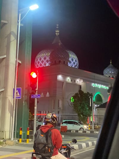 This media file captures a scene at night on a street in Jawa, Indonesia. The primary subjects are a mosque and a person on a motorcycle. The mosque features prominent domes with a patterned design, illuminated by colorful lights, suggesting it is a place of worship. The building also has Arabic script, rendered in neon green, above an arched entryway.
In the foreground, a person wearing a red jacket and a decorated helmet is seated on a motorcycle, facing away from the camera. They are carrying a black backpack. The motorcycle is positioned on the side of the road, near a traffic light that is currently red, indicating a stop.
The setting appears to be urban, with streetlights and other illuminated signs visible in the background. A white car is also seen on the road. The ground is paved, with striped road markings and bollards separating lanes or areas. The overall atmosphere is nocturnal and illuminated, possibly a busy intersection or a well-lit area near the mosque. The time of day is clearly night, and the weather appears to be clear as there are no indications of rain or fog. The image is taken from a slightly elevated perspective, likely from another vehicle or a higher vantage point on the street. There are no explicit emotional cues, but the vibrant lighting suggests activity and possibly a festive or welcoming ambiance around the mosque.