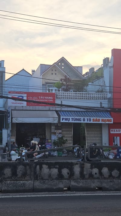 The image captures a street scene in Cam Đức, Vietnam, featuring storefronts along a roadway. The building on the left has the TotalEnergies sign and a red sign above it that says CỬA HÀNG SÁU HẠNG, which translates to Sau Hang Shop, specializing in the sale of spare parts. To the right, there is another storefront with a blue awning, reading PHỤ TÙNG Ô TÔ SÁU HẠNG, meaning Sau Hang Auto Parts. Further to the right is a HEAD store. Power lines are visible against a pale sky in the background. People are present on the sidewalk in front of the stores, interacting in the foreground. Parked scooters line the sidewalk. A gray concrete wall borders the right edge of the road.