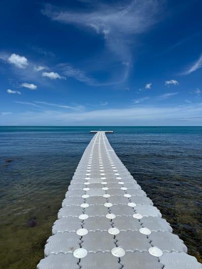 A floating walkway extends from the shore into the ocean under a bright blue sky with scattered white clouds. The walkway is made of modular, grey plastic units with white circular accents. The ocean water is a clear turquoise near the shore, becoming a deeper blue further out. Rocks are visible beneath the surface of the water on the left side of the frame. In the distance, two figures can be seen standing on a small structure in the water. The scene is located in Choeng Mon, Thailand.