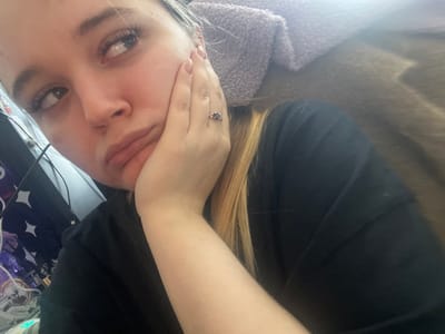 A female individual is depicted in a close-up, indoor setting within Pushchino, Russia. The subject has light skin, prominent eyelashes, and light-colored hair with visible blonde streaks. She is wearing a dark, short-sleeved garment. Her right hand, adorned with a ring featuring a central dark stone flanked by smaller stones, supports her chin, with her fingernails exhibiting a light-toned polish. Her facial expression presents with downturned lips and eyes directed upwards and to the right. The immediate background on the left side includes a partial view of a dark, vertical object, likely an electronic device, with faint star-like reflections. The background on the right side consists of blurred, textured fabrics, specifically a light purple material and a darker brown material, consistent with soft furnishings in a domestic environment. No specific external landmarks or architectural features are visible to denote a precise street address or specific building within Pushchino.