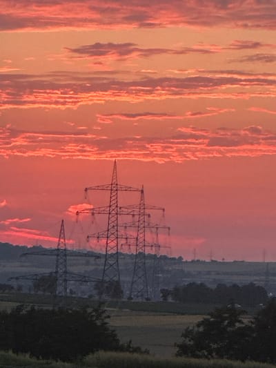The image depicts a vibrant sunset over a rural landscape, likely near Ludwigsburg, Germany, given the characteristic high-voltage power pylons visible. The sky is a dramatic display of orange and red hues, with layered clouds catching the last rays of the sun. In the foreground, silhouetted against the colorful sky, are several large, lattice-style power pylons. These pylons are arranged in a line, with the closest one being the most prominent. The landscape below the pylons consists of rolling hills, fields, and scattered trees, all rendered in dark, muted tones due to the fading light. The overall scene conveys a sense of industrial infrastructure set against the natural beauty of a sunset. There are no people or animals visible in the image.