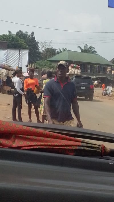 The image depicts a daytime street scene in Igbo Ukwu, Nigeria, observed from within a vehicle.

In the immediate foreground, the top of a dark vehicle dashboard or door sill is visible, overlaid with a folded umbrella or fabric displaying a red and black pattern with circular gold motifs.

The midground features several individuals. A male in a dark blue polo shirt with a red collar and khaki shorts, wearing a camouflage baseball cap, walks from left to right, looking towards the left. To his left, a group of people includes a female in a white long-sleeved top and dark pants with "SP" lettering, standing still. Beside her, another female in a bright orange short-sleeved top and dark skirt holds a yellow bucket and a dark bag. Behind them, a figure with items balanced on their head is visible, along with another person in a light-colored patterned dress.

On the unpaved and partially paved road behind the individuals, a dark-colored SUV with bundles strapped to its roof rack is either parked or moving slowly. Further down the road to the right, a tricycle (Keke Napep) and a white motorcycle are present.

The background consists of structures and vegetation. To the far left, a market stall covered with white sheeting is visible. Beyond this, buildings with distinctive green corrugated iron roofs are interspersed with abundant green foliage, including palm trees. Overhead power lines stretch across a light grey, overcast sky. The overall environment suggests a busy commercial or residential area.