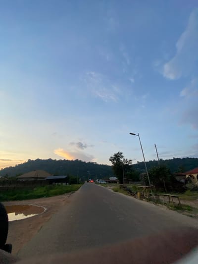 This image presents a twilight view of a road in Uffah, Nigeria. A dark grey asphalt road extends from the foreground into the distance, flanked by reddish-brown dirt shoulders. In the lower left foreground, a large, irregularly shaped puddle of water reflects the sky's hues. A dark, undefined object is partially visible in the extreme lower left.
The midground features dense green foliage and scattered residential structures, including houses with brown and dark grey sloped roofs. On the right side of the road, a tall, dark streetlight pole stands, along with a wooden utility pole. Further along the right shoulder, two rustic wooden structures resembling tables or stalls are visible, with a yellow container placed near one.
In the distance, at least two vehicles with illuminated red taillights are present on the road, with a white vehicle also discernible on the right side. The background is dominated by a range of dark, tree-covered hills or mountains, featuring some visible rocky outcrops. The sky overhead transitions from a light blue at the zenith to a pale yellow and orange gradient near the left horizon, populated by sparse, wispy white and grey clouds. One elongated cloud above the left hillside exhibits a prominent orange glow, indicative of a low sun.