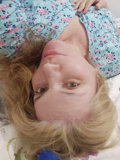 The image shows a close-up, upside-down view of a Caucasian woman lying on her back. She has fair skin, light brown and blonde hair, and is wearing a light blue, floral-patterned shirt. Her eyes are open and looking towards the camera. Her left hand is resting on her chest, near her shoulder. The background appears to be a bed with a patterned sheet. The lighting is soft and natural. There is no visible indication of the city of Al Mas'oudi, United Arab Emirates in the image.