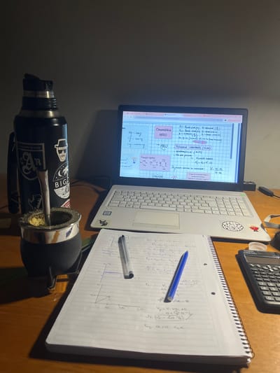 On a wooden desk, a laptop displays notes on kinematics (MRU), next to a notebook filled with handwritten physics calculations. A calculator sits nearby. In the foreground is a mate gourd with a bombilla, and a thermos adorned with stickers, including one that reads "Argentino de Rugby." One sticker depicts a design including the word "BIGO" and a cartoon figure. Based on the sticker "Argentino de Rugby" and the term "Argentino" and other indications on the screen and thermos, the location may be Gerli, Argentina, a city within Argentina. The overall environment appears to be a study or workspace, with a focus on academic pursuits.