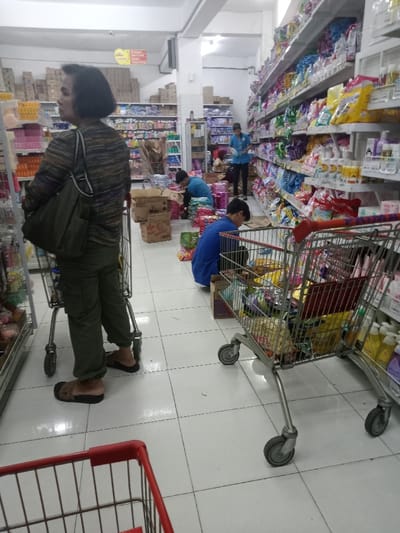 The image depicts an indoor retail environment, specifically a supermarket aisle, located in Nganjuk, Indonesia. The scene is illuminated by overhead fluorescent lighting, reflecting off a white tiled floor and white ceiling.
In the foreground, slightly to the left, a person with short, dark hair, wearing a horizontal striped long-sleeved shirt, olive green cargo pants, and brown slip-on sandals, stands with their back mostly to the camera, facing left. A dark green shoulder bag is worn over their left shoulder. A chrome shopping cart, largely empty, is positioned directly beside them.
In the mid-ground, towards the right, two individuals wearing blue short-sleeved shirts are observed crouching on the floor. They are surrounded by open and closed cardboard boxes and numerous colorful packaged products, indicative of merchandise stocking or sorting activity. One of these crouching individuals is positioned near a second chrome shopping cart, which is visibly filled with various packaged goods. Behind these two individuals, a third person, dressed in a light blue collared shirt and dark pants, stands upright, appearing to supervise or engage in store operations near the stocked shelves.
The background features long aisles with multi-tiered shelving units densely packed with a wide assortment of consumer products, including toiletries, snacks, and household items. Another red-handled shopping cart is partially visible in the extreme lower foreground.