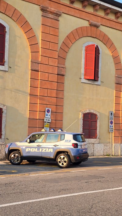 This media file displays a police car parked on a street next to a building with a distinctive architectural style. The building has a yellow facade with arched brickwork and red-shuttered windows. The police car, labeled "POLIZIA" and "113" in Italian, is a white SUV with blue accents and markings. It appears to be a Jeep Renegade. The street has asphalt pavement with painted white and yellow lines.

Several traffic signs are visible. One sign indicates a parking restriction with a 24-hour duration. Another sign features a symbol for a police station or service area. The scene is illuminated by the warm, golden light of either sunrise or sunset, casting long shadows. The weather appears clear and pleasant.

The image is captured from a high angle, looking down on the scene. The perspective suggests the photographer might be on an elevated position, perhaps an upper floor of a building or a nearby elevated area. The orientation of the image is rotated, with the street running vertically.

The specific location is identified as Peschiera del Garda, Italy, which is a town known for its historical fortifications and scenic beauty along the southeastern shore of Lake Garda. This context suggests the image might have been taken in a town center or a residential area with such architectural features. No people are visible in the frame, focusing the attention on the car and the building. The overall mood is tranquil, with the sunlight adding a sense of warmth and calm to the scene.