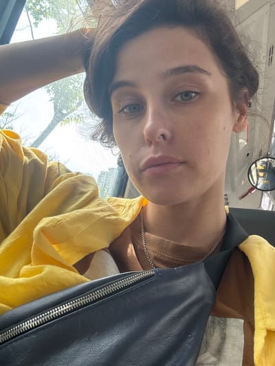 A young woman with dark hair is taking a selfie in a vehicle, possibly a bus, in Busan, South Korea. She is wearing a brown t-shirt and a bright yellow shirt over it. A dark blue fanny pack is slung across her chest, and a silver necklace is visible. Her left arm is resting on her head. The background shows a window with trees and buildings visible outside. A circular mirror, likely from the vehicle, is positioned to the right of her face, reflecting parts of the interior, including a red strap. The lighting suggests it is daytime, with sunlight streaming through the window.