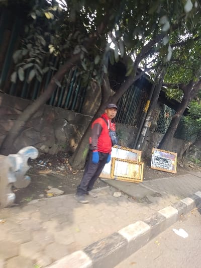 A man in a red long-sleeved shirt and blue gloves stands next to a stone wall along a concrete sidewalk in Kecamatan Cimahi Selatan, Indonesia. Three framed pictures lie on the ground to the right of the man. The pictures appear to have gold trim and are covered with glass. There is a large tree behind the man. The sidewalk curves to the right of the man. There is a section of pavement to the right of the sidewalk. There is a white object on the pavement. The man is facing right, looking towards the pictures on the ground. The scene appears to be a street corner. The ground is covered in dirt and debris. The sky is cloudy. The coordinates -6.901, 107.563 correspond to a location in Kecamatan Cimahi Selatan, Indonesia.