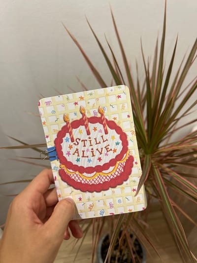 A person's hand holds a rectangular birthday card in front of a Dracaena plant. The card has a yellow checkered background with colorful confetti and stars. A drawing of a red-frosted cake with three lit candles is on the front of the card. The words "STILL ALIVE" are written on the cake in a festive font. A blue binder clip is attached to the left edge of the card. The background is a plain, light-colored wall and the plant's foliage extends into the frame. The image was taken in Chang Phueak, Thailand.