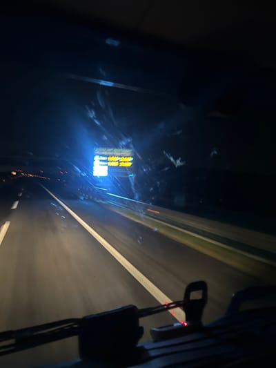 The image depicts a nighttime scene from the interior of a moving vehicle, likely a truck, traveling on a multi-lane highway. The perspective is forward-facing through the windshield, which exhibits glare and streaks, possibly from dirt or reflections. The location is the A14 highway near Cerignola, Italy, indicated by a prominent overhead digital variable message sign displaying "MOLTO TRAFFICO" (heavy traffic) and "A14" in bright blue and yellow text, alongside numerical values such as "1:00" and "2:00," likely denoting travel times. The highway pavement is dark, with white solid and dashed lane markings visible, streaked due to motion blur. Distant red taillights of other vehicles are visible on the left side of the highway, along with fainter white lights. In the immediate foreground, the dark silhouette of the vehicle's dashboard is visible, including what appear to be control stalks or wiper arms with small, illuminated red indicator lights. No individuals are directly visible.