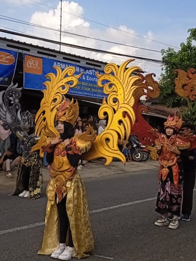 The image captures a street scene in Winong, Indonesia. Three individuals are costumed in ornate, colorfully embellished garments reminiscent of traditional Indonesian attire. The costumes feature prominent wing-like backpieces: one in black and silver, another in gold, and a third in red and gold. Each individual is wearing distinct headwear. The person in the gold costume is wearing a hijab. The other two are wearing matching shoes with thick soles.

In the background, a group of spectators is visible, along with a parked motorcycle. Overhead, electrical wires are strung. A blue banner displays text reading "ARMAM PLASTIC," suggesting a local business, and includes the phrase "(Sebelah Barat KUA Winong, Desa Pekalong.n)".