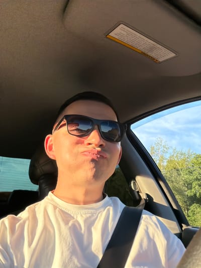 An individual is captured in a close-up selfie or portrait while seated inside a vehicle, identified as a car in Yus'ki, Russia. The person, appearing to be male, is seen from the chest up, with their head tilted to the right. They are wearing dark sunglasses and a white t-shirt, secured by a seatbelt across their chest. Their lips are pursed, suggesting a playful or affectionate expression.
The setting is bright and sunny, indicated by clear blue skies with scattered clouds visible through what appears to be a sunroof or open window. Green foliage from trees can be seen outside the car, suggesting a natural outdoor environment. Golden sunlight illuminates the person's face and shirt, hinting at either early morning or late afternoon light.
Visible within the car's interior is a sun visor with a prominent "WARNING" label. Despite some text being obscured or garbled by the image quality and angle, key phrases like "DEATH OR SERIOUS INJURY can occur" are discernible. The label also mentions "SAFEST place for children," "airbag," "seat in the front," and "SEAT BELTS and CHILD RESTRAINTS," advising to refer to a "manual for further information." The image is rotated, giving a unique perspective to the scene.