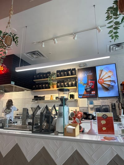 Here's a detailed description of the image:
**Setting:** The image appears to be taken inside a Lukumades dessert shop. The setting has a modern aesthetic with white walls, herringbone tile details, black tiled accents, and track lighting. A neon sign is partially visible near the lower edge of the image.
**Objects and Elements:**
* **Signage:** Digital displays advertise Lukumades gelato and the "Junior Pack" featuring donuts and drinks. Printed menus and signage are visible near the counter.
* **Products:** Packaged goods are stocked on shelves.
* **Equipment:** A coffee grinder, espresso machine, and other food preparation equipment are situated on the counter.
* **Accessories:** Mugs, napkins, and serving utensils are arranged on the counter space.
* **Decor:** Plants in hanging baskets adorn the interior.
**People:**
* An employee, wearing a "Lukumades" branded shirt, is visible working behind the counter.
**Action and Interactions:**
* The employee is likely engaged in preparing or serving food or drinks.
**Inferences:** The setting suggests a food service business catering to a modern and possibly family-oriented clientele. The presence of various equipment and products indicates a functioning retail environment.
**Location:** Based on the store name, it is likely that this photo was taken in Jacksonville Beach, FL, United States of America.