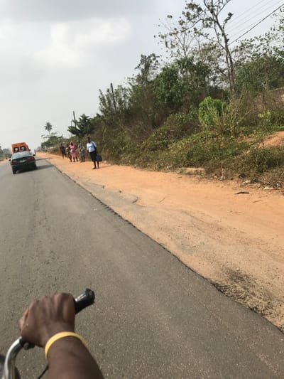 The image depicts a roadside scene in Ogbese, Nigeria, from the perspective of an individual riding a bicycle or motorcycle, evidenced by a dark-skinned right hand gripping a black handlebar in the lower foreground, wearing a yellow wristband. An asphalt road, exhibiting cracks and uneven edges, occupies the left and central portions of the frame, alongside a wide, reddish-brown dirt shoulder to its right.

In the mid-ground, five individuals are visible on the dirt shoulder. The foremost person is a woman in a light blue top and dark skirt, carrying a dark bag, oriented to her left. Behind her, four additional individuals, primarily women in casual clothing, stand further down the road.

A dark sedan travels away from the viewer on the asphalt road in the mid-ground. Behind it, a light-colored vehicle with an orange or yellow roof attachment and red/white markings on its rear is partially visible, also moving away.

The environment features dense green and brown vegetation, including bushes, trees, and some bare-branched trees, lining the right side of the road. Power lines are visible running through the foliage. A single palm tree stands in the distant left. The sky overhead is overcast and light gray.