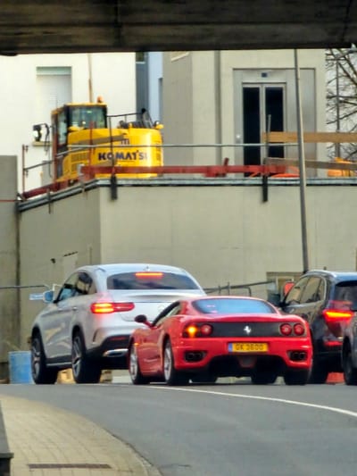 An urban street scene in Schifflange, Luxembourg, is depicted from under an overhead concrete or metal structure. Three vehicles are positioned on a paved road, facing away from the viewer. The central vehicle is a red Ferrari 360 Modena, displaying Luxembourg license plate "GX 3600" and illuminated taillights. To its left and slightly behind is a silver Mercedes-Benz GLE Coupe, also with illuminated taillights. A dark-colored SUV is partially visible on the far right, behind the Ferrari, showing its left rear and illuminated taillights. A grey paved sidewalk borders the left side of the road, and a blue barrel is partially visible near the sidewalk on the left. In the background, on an elevated platform or building roof, a large yellow Komatsu excavator is visible, surrounded by red and silver construction barriers. Behind the excavator, several light-colored buildings are present, one featuring a prominent external elevator shaft with dark doors and another with a white window blind. No individuals are visible in the scene.