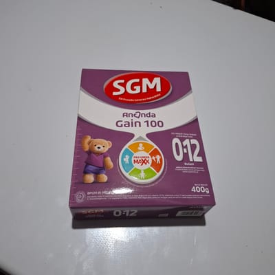 The image shows a close-up, slightly angled view of a SGM Ananda Gain 100 formula milk box, predominantly purple and white, resting on a plain, light-colored surface, possibly a table. The box features branding, product information, and graphics, including a teddy bear illustration and a circular graphic with icons. The lighting is soft and diffused, casting minimal shadows. The background is out of focus, showing a blurred dark area at the top right, suggesting an indoor setting. There are no people visible in the image, and no specific location in Reudeuep, Indonesia can be determined from the visual information provided.