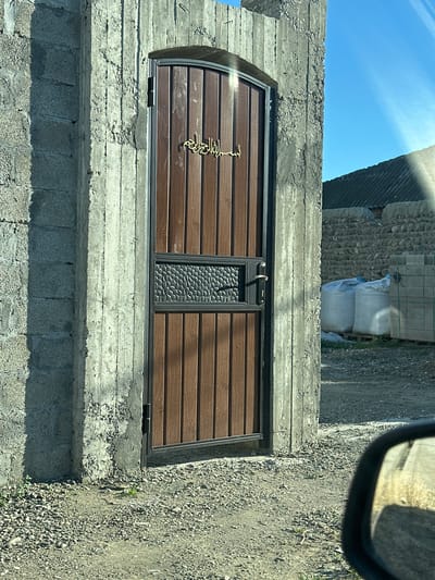 The image shows a closed wooden gate with an arched top, set into a concrete structure. The gate has a decorative metal insert in the upper section and a gold-colored Arabic inscription on the upper part of the wooden planks. The concrete structure appears to be part of a wall or entryway, with a rough, unfinished texture. To the right of the gate, a building with a dark, sloped roof is visible, constructed with stone or brick. Several large white sacks and stacks of grey building blocks are also present in the background. The ground is covered with gravel and dirt. The sky is blue and clear, with some sunlight creating lens flares. This scene is located in the city of Miatli, Russia.