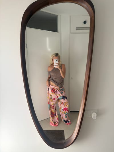 A blonde woman is taking a mirror selfie in a room with white walls. She is wearing an off-the-shoulder brown top and pink pajama pants with a colorful leopard print. Her left hand is on her hip, and her right hand is holding a smartphone. The room appears to be a hallway or entryway, with a closed door in the background and a light switch and a small white device on the wall to the right. The mirror itself is large and has a dark wooden frame with an irregular shape. The overall setting is modern and minimalist.