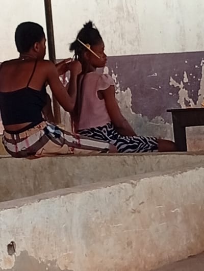 Within a room in Omicha Umu Awada, Nigeria, two females are observed. An adult female, characterized by short dark hair and dressed in a black spaghetti strap crop top paired with horizontally striped trousers (white, brown, and black), is actively braiding the hair of a younger female. The younger female, wearing a light pink short-sleeved top and black and white zebra-print trousers, presents dark hair with a yellow comb inserted on the right side of her head. Both individuals are seated on a multi-tiered, light-colored concrete or stone structure with a rough surface texture. The background wall exhibits extensive paint peeling, exposing a darker grey-purple layer beneath a lighter top coat. A dark-colored table or similar furniture item is partially visible on the far right, and a light-colored vertical support beam is partially visible on the far left.