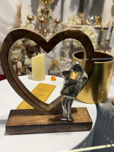 The image shows a close-up, eye-level, indoor shot of a decorative item. The main subject is a heart-shaped wooden frame with a small, silver figurine of a person with their arms raised in front of them, positioned on a wooden base. The figurine is inside the heart frame. Behind the figurine, to the right, is a cylindrical gold-colored container. To the left of the figurine and inside the heart, is a pale yellow, cylindrical candle. A yellow tag with text is placed on the surface in front of the wooden base. The background is blurred, showing hints of decorative elements like flowers and metallic objects, suggesting a retail or display setting. The scene does not provide any information that would allow for determining its location in Ilidža, Bosnia and Herzegovina.