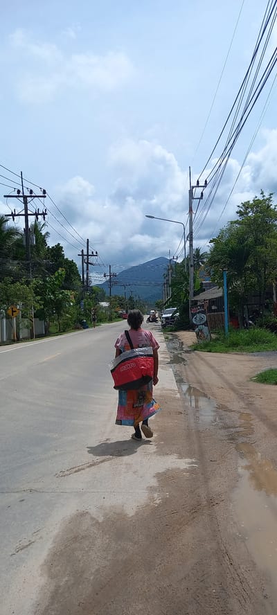 An elderly person wearing a red plastic poncho walks on the right side of a two-lane paved road in Thong Sala, Thailand. The road has patches of standing water along the curb. Power lines and utility poles run along the left side of the road. Lush green trees and vegetation flank both sides of the road. In the distance, a mountain is visible. A few buildings are interspersed among the trees. The sky is blue with scattered clouds.