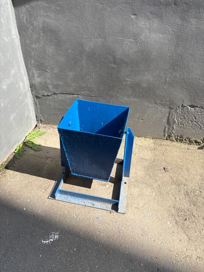 The image shows a blue metal trash receptacle on a concrete surface, likely an alleyway or courtyard, with two grey concrete walls forming a corner. Sunlight casts distinct shadows on the ground and the receptacle. A few green weeds are growing near the base of the left wall. The scene is located in Blagoveshchensk, Russia.