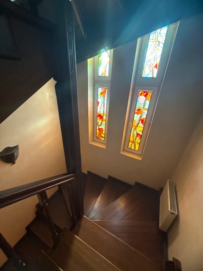 The image shows a view looking down a dark wooden spiral staircase. The staircase has a wooden railing with carved posts. Two narrow, vertical stained-glass windows are visible at the top of the staircase, letting in colorful light. One window shows an abstract design with yellow, red, and blue glass, and the other shows a vine with orange and yellow leaves and red flowers. To the left of the staircase, a dark sconce is mounted on a cream-colored wall. A white radiator is mounted on the wall to the right of the bottom of the staircase. The environment appears to be an indoor staircase in a residential or public building in the city of Yukki, Russia. There are no people visible in the image.
