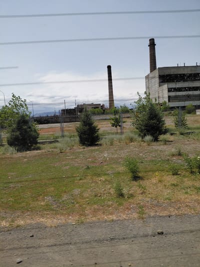 The image captures a view of an industrial area under a cloudy sky. Dominating the scene are two large, brick chimneys, one taller than the other, situated next to a large, rectangular, concrete industrial building with broken windows. The building appears weathered and abandoned. In the foreground, there is a patch of dry, brown grass interspersed with green patches and scattered stones, indicating a dry, possibly arid environment. Several small, conical evergreen trees and other sparse vegetation are scattered throughout the area, some behind a metal fence. The overall impression is one of an abandoned industrial site, likely in a somewhat remote or neglected part of a city. The geographical context provided by the prompt suggests this is an industrial facility in Бұлақты, Kazakhstan.