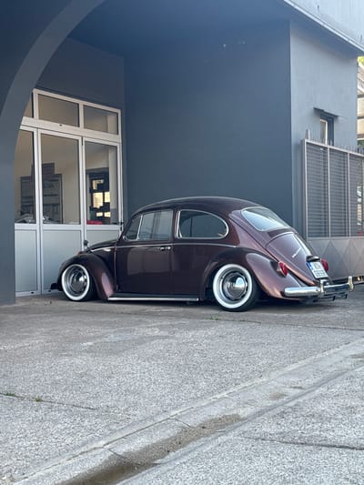 A dark metallic brown, lowered Volkswagen Beetle is parked on a concrete surface in front of a dark gray building. The building has a large glass door and window section on the left, revealing an interior with shelves and tools. To the right of the building is a metal grate fence. The car has whitewall tires and chrome hubcaps. The license plate on the rear of the car reads "K DV G996B". The scene appears to be in an industrial or commercial area. The time of day appears to be daylight, with shadows indicating the sun is to the right. The overall impression is one of a well-maintained, classic car in a somewhat utilitarian setting.