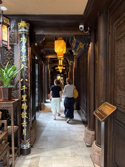 This image captures a traditional Chinese architectural corridor, likely part of a temple or cultural site, located in Qingyang District, China. The corridor features dark brown wooden pillars, overhead beams, and meticulously carved wall panels.

In the foreground on the left, a tall, dark wooden pillar is prominently displayed, featuring vertical gold Chinese characters that read, from top to bottom, "功名生外千里路披雲月" (Gōngmíng shēng wài qiānlǐ lù pī yún yuè). The pillar is ornate, with a gold and silver decorative finial and a red and gold patterned base. To its left, a potted plant with long green leaves rests on a dark wooden carved stand. The left side of the corridor consists of dark wooden screens with lattice patterns, some partially open.

Two women are seen walking away from the camera down the length of the corridor on a light grey tiled floor. The woman on the left wears a black short-sleeved top, light-colored (possibly white or beige) cargo pants, and sandals. The woman on the right wears a white long-sleeved top, dark blue or black pants, white sneakers, and carries a bright yellow shoulder bag on her left shoulder. Faint figures are discernible further in the background.

Overhead, several square traditional Chinese lanterns with illuminated yellow panels depicting painted scenes hang from the dark wooden ceiling. Modern white recessed light fixtures are also visible in the ceiling. A white security camera is mounted on an overhead wooden beam on the right side, connected by a white cable. On the right, dark wooden carved panels line the wall. In the right foreground, a brown, angled informational sign with white Chinese text is positioned on a stand. The title of the text reads "千年廊桥记始见" (Qiānnián lángqiáo jì shǐjiàn), followed by smaller descriptive Chinese characters. The bases of the wooden pillars throughout the corridor are supported by carved stone pedestals with intricate patterns.