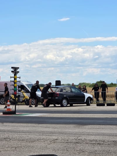 The image depicts a drag racing event under a bright, partly cloudy sky, possibly in Akatsievo, Bulgaria. In the foreground, a grey asphalt track displays tire marks. To the left, a drag racing tree with illuminated lights stands near an orange traffic cone and a spare tire. Behind the tree, a yellow scooter and a white van are parked. Two men in black shirts and shorts are pushing the rear of a black and white car, with one man's hands on the trunk and the other on the rear bumper. A second black car is partially visible to the right. In the background, a line of people stands behind a metal fence, watching the proceedings. The scene suggests a warm, outdoor event with a focus on automotive competition.