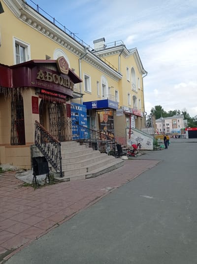 The image captures a street scene in Degtyarsk, Russia, focusing on the facade of a building housing several businesses. The building is painted a light yellow with white architectural detailing along the roofline and around the windows. A prominent burgundy awning with the gold lettering "АБСОЛЮТ" (ABSOLUTE) and "ГОРОДСКОЙ ЦЕНТР НЕДВИЖИМОСТИ" (CITY REAL ESTATE CENTER) is visible on the left. Below this awning, an entrance is protected by ornate black metal gates. To the right of this entrance, a set of wide, light-colored tiled stairs with a black metal railing leads up to another entrance.
Further to the right, a blue sign with white lettering reads "АПТЕКА 66" (PHARMACY 66), indicating a pharmacy. Next to it, a sign advertises "ТОРТЫ" (CAKES) with images of cakes and pastries. More businesses are visible further down the street, with people walking in the background. The street in the foreground is paved with red and gray tiles and asphalt. A red stroller is parked near the stairs, and a trash bin is located at the bottom of the stairs. The sky above is partly cloudy.