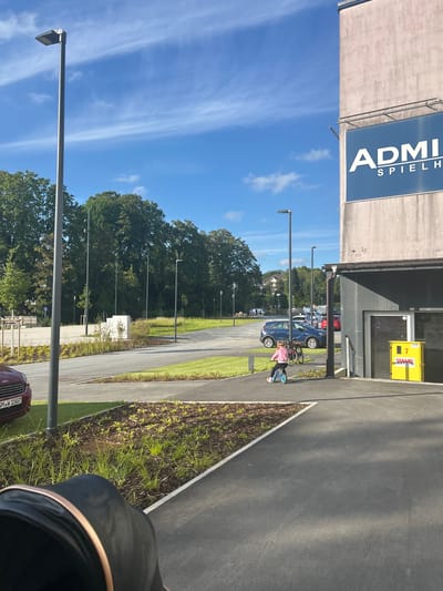 A little girl wearing a pink top and leggings rides a small blue balance bike on a paved path. She is on the right side of the image, moving from right to left. Behind her, a bicycle is parked. A concrete building with a large blue sign that reads "ADMI SPIELH" is visible on the right. A yellow utility box with the number "7" and "WWB" printed on it is positioned next to the building's entrance. The background includes a parking lot with several cars, trees, and a bright blue sky with scattered white clouds. The scene appears to be in Waldbröl, Germany, with a park-like setting adjacent to a commercial or recreational building.