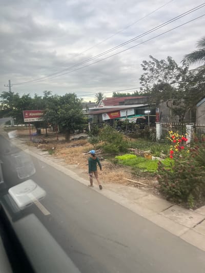 The image presents an outdoor roadside scene in Ninh Tây, Vietnam, under a heavily overcast sky. A paved asphalt road occupies the immediate foreground, exhibiting reflections on its surface. Adjacent to the road, a concrete sidewalk runs parallel.
A single individual is observed walking on the sidewalk, moving away from the viewer towards the background. The person is dressed in a blue cap, a dark green long-sleeved shirt, tan trousers, and light-colored footwear. They carry a white rectangular object, possibly a small bag or container, in their right hand, held at their side.
Beyond the sidewalk, a strip of ground features dry, light brown vegetation and sparse debris. Further back on the left, a red billboard displays the "Havoline" logo and the Vietnamese text "SUA XE TRU," positioned near a utility pole. Green trees with dense foliage are interspersed across the midground.
To the right of the dry vegetation, cultivated patches of green plants, appearing to be small crops or garden beds, are visible. These are situated in front of a property enclosed by a dark fence. Within this property, several buildings are present, including one with a red roof that also features a "Havoline" sign with Vietnamese text, suggesting a repair establishment. Faint outlines of parked vehicles, likely motorcycles, are discernible near this building. On the far right, a bush with red and yellow flowers is present. Multiple power lines traverse the grey sky overhead.