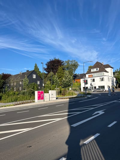 This image depicts a street scene in Waldbröl, Germany, under a bright blue sky with wispy white clouds. In the foreground, a two-lane road is visible with white lane markings, including a left-turn arrow. A shadow of a person and a fence is cast across the road from the left.
On the left side of the street, a dark-colored, multi-story building with multiple windows is surrounded by lush green foliage and trees. A pink and white advertisement display stands on the sidewalk in front of this building.
To the right of the road, a white, multi-story building with a brown tiled roof stands. This building appears to be a commercial establishment, with a sign visible above the ground-floor windows. In front of this building, a silver car is parked.
Two pedestrians are crossing the street on a marked crosswalk in front of the white building. One pedestrian is wearing a light-colored shirt and dark pants, while the other is wearing a dark top and shorts. Traffic lights are visible at the intersection, with one light showing red. The environment is characterized by a mix of residential and commercial buildings, mature trees, and well-maintained landscaping. The overall atmosphere suggests a sunny day in a suburban or small-town setting.