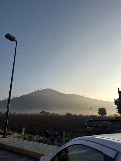 The image shows an early morning scene in San Andrés Payuca, Mexico. The sky transitions from a clear blue to a hazy, light yellow near the horizon, suggesting sunrise. A long mountain range, lightly forested along its ridge, runs diagonally through the center of the frame. A row of cultivated fields lies adjacent to the mountain, possibly a farm. Two people are visible on the right side of the frame, apparently working along the edge of the fields. The corner of a white car intrudes into the frame from the upper right. A street light is visible in the lower left corner. A concrete curb is adjacent to the cultivated fields.