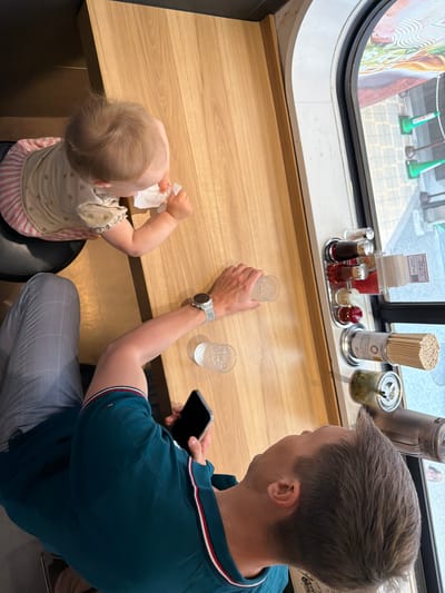 An overhead shot depicts an adult male and an infant seated at a light-wood counter in a casual Japanese eatery, consistent with a location in Osaka, Japan. The adult male, positioned on the right, wears a dark teal polo shirt with red, white, and blue trim on the collar and sleeves, and light grey checkered trousers. His left arm rests on the counter, revealing a light grey smartwatch on his wrist, while his right hand holds a black smartphone. A clear glass with ice and water is positioned in front of him.

To his left, an infant is seated on a black high chair or booster seat, facing the counter. The infant has light brown hair with a distinct swirl on the crown and wears a light-colored top with faint heart patterns and a pink and white striped lower garment. Both of the infant's hands are occupied with a crumpled white tissue or napkin.

The counter features a polished light-wood surface. Along the edge nearest the window, a metal tray holds various seasonings: two bottles with dark and reddish liquids, two smaller containers with yellow and red pastes, and a clear shaker. Adjacent to this, a tall metal cylinder contains numerous long, uncolored sticks, likely dried noodles or chopsticks. Another clear glass jar filled with green pickled items is visible further down the counter.

Through a large window to the right, a vibrant advertisement for food (possibly ramen) is partially visible. The exterior scene shows a concrete sidewalk or street with two green traffic cones standing upright. The lighting suggests daytime.