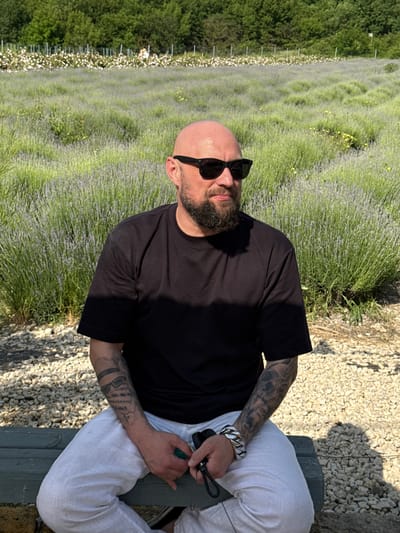 A bald, bearded man wearing sunglasses and a black t-shirt sits on a green bench in front of a lavender field. The man is positioned in the center of the frame, with his body angled slightly to the right. His left arm, adorned with tattoos, rests on his lap, while his right hand holds a green object. The background features a vast expanse of purple lavender flowers stretching towards a line of trees and vegetation. The ground in the foreground is covered with small, light-colored stones. The lighting suggests it is daytime, with sunlight casting shadows.