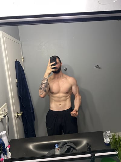 A man is posing in a mirror in what appears to be a bathroom or dressing room, shirtless and wearing black shorts. He is holding a cell phone in his left hand, partially obscuring his face, and is flexing his right arm. The man has tattoos on his left arm and a beard. The room has gray walls, a towel hanging near the bottom of the frame, and a black vanity or cabinet to the right, with items on top, including what appears to be a plant and some toiletries. There is a mirror frame visible on the left and right edges of the image. There is a light switch at the bottom of the mirror. The location of the photograph is a bathroom in Arlington Heights, Illinois, United States of America.