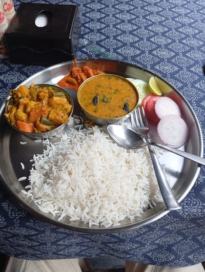 This close-up shot captures a traditional Indian thali meal served on a round, stainless steel plate, set on a blue tablecloth with a light-colored, repeating ornamental pattern.

The main subject is the food itself. A large mound of white, fluffy basmati rice dominates the lower left side of the plate. Two small, round metal bowls hold different dishes: the left bowl contains a vibrant mixed vegetable curry with potatoes, carrots, green beans, and cauliflower in a rich, orange-hued gravy, while the central bowl holds a yellow lentil soup (dal) garnished with what appears to be black mustard seeds and curry leaves. A small dollop of reddish-orange chutney or pickle is visible above the dal.

Accompanying the main dishes is a fresh salad on the right side of the plate, consisting of thinly sliced red onions, tomato slices, and a piece of cucumber, with a wedge of lime for squeezing. A metal fork and spoon are neatly arranged on the plate, resting on the rice and salad.

In the upper left background, a black, rectangular napkin dispenser is partially visible. Behind the dispenser, fragmented text from a label or menu can be seen, including "Catch Up," "SPRINKLE," and "Table."

The scene depicts a mealtime setting, likely in a restaurant or eatery, as suggested by the presentation and the napkin dispenser. The overall impression is that of a simple, wholesome Indian meal ready to be enjoyed in Arambol, India.