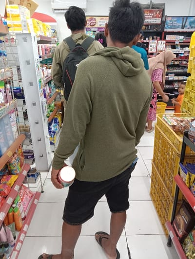 The image shows a man in a green sweatshirt and black shorts standing in a store aisle. He is holding a white cylindrical can in his right hand. A man wearing a grey t-shirt and a black backpack is standing in front of him. There is a woman in a pink shirt to the right of the man in the green sweatshirt and a woman in a brown and pink shirt to the left. There is a child in a yellow shirt to the right of the man in the green sweatshirt. The shelves in the store are stocked with various items. The floor is tiled and white. The location of the store is at (latitude: -6.953, longitude: 110.239) in the city of Kaliwungu, Indonesia. The photo was likely taken with a smartphone camera. The image is of low resolution.