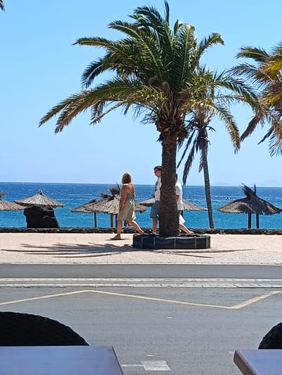 The image presents a vibrant coastal scene in Puerto Del Carmen, Spain, bathed in bright midday sunlight. A clear, deep blue sky meets the equally blue ocean, which stretches across the horizon. A paved promenade runs along the coastline, offering views of the sea.
Dominating the scene are several tall palm trees with green fronds, casting long, sharp shadows on the paved walkway, indicative of the strong sun. A large palm tree stands prominently in the mid-ground, its base enclosed by a dark, circular planter.
Along the water's edge, several rustic straw beach umbrellas are visible, with a dark, rocky formation nestled in the ocean near one of them. The tranquil blue waters show minimal choppiness.
Three individuals are captured walking along the promenade. A woman in a light-colored, possibly beige or light green, strapless dress walks towards the left, her back mostly to the viewer. Behind the central palm tree, a man in a white t-shirt and shorts is visible, followed by another person, also wearing a white t-shirt, suggesting a casual stroll.
In the immediate foreground, parts of a dark, textured object, possibly a chair or a car tire, and a light-colored table surface are visible, implying the photograph was taken from a seated position at an outdoor establishment. Below the promenade, a road or street with a yellow marking line is visible. The overall atmosphere is bright, relaxed, and typical of a sunny holiday destination.
