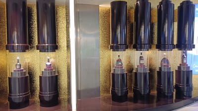 The image shows a display of liquor bottles inside cylindrical glass cases, each standing on a tiered, dark-colored pedestal. Above each glass case is a dark, cylindrical structure. The background is a textured gold wall. On the left side of the image, there are two such displays. On the right side, there are four more displays, with the rightmost one partially obscured by a blue sign that reads "MAX" at the top and "ENJOY" at the bottom. The scene is likely in a retail or commercial establishment, possibly a duty-free shop or a high-end liquor store. The presence of the "MAX" sign suggests a connection to the company "PT Maxima Global Prima" which operates duty-free shops in Indonesia, including at the Soekarno-Hatta International Airport, a key transit hub serving Tangerang and the greater Jakarta metropolitan area.