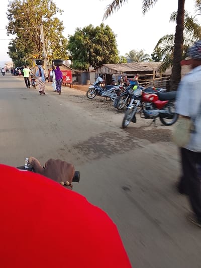 A street scene in Ube, Nigeria, is captured from a low, first-person perspective, likely from a person riding a bicycle. The immediate foreground shows a right hand, adorned with a beaded bracelet, gripping a black bicycle handlebar, with a vibrant red garment obscuring the lower-left frame. The unpaved road, displaying tire tracks and scattered dark, wet patches, extends into the midground.
On the left side of the road, four women walk away from the viewer; three are carrying large, dark basins balanced on their heads. They wear patterned traditional attire, including head coverings. A man in a light green shirt and dark trousers walks beside them, pushing a bicycle. Additional pedestrians are visible further down the road. On the right side, a blurred figure, possibly a male, partially obscured, is in motion.
Numerous motorcycles are parked or moving along the right edge of the road, including a prominent red motorcycle with chrome accents and a lighter-colored scooter. Behind these vehicles, a row of rustic, single-story structures with corrugated metal roofs and wooden supports, suggestive of market stalls or small shops, lines the roadside. Some structures display red signage. Tall green trees, including palm trees, are interspersed throughout the background under a bright, clear sky. Power poles with overhead lines are visible on the left side of the road.