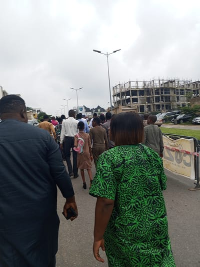 A group of people are walking on a paved street in Guzape, Nigeria, with a building under construction in the background. The sky is overcast. The people are dressed in various casual and formal attire. In the foreground, a man in a dark blue agbada is holding a mobile phone in his right hand. To his right, a woman in a green and black patterned dress is walking away from the camera. Behind them, a line of individuals, including men, women, and a child with a pink backpack, are also walking along the street. The construction site features exposed rebar and concrete structures. Vehicles are parked on the side of the road. A white barrier with black text and red tape is visible to the right of the frame.