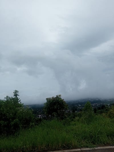 The image presents an elevated landscape view of the city of Babaliya Mal., India, under heavy overcast conditions. The immediate foreground shows a grassy embankment with a narrow strip of paved road, marked by a white line, at the bottom. The midground is filled with dense green foliage and trees. In the distant background, a sprawling urban settlement, consistent with Babaliya Mal., is visible through atmospheric haze and low-hanging clouds that partially obscure the horizon. No individuals, animals, or specific actions are discernible within the frame.